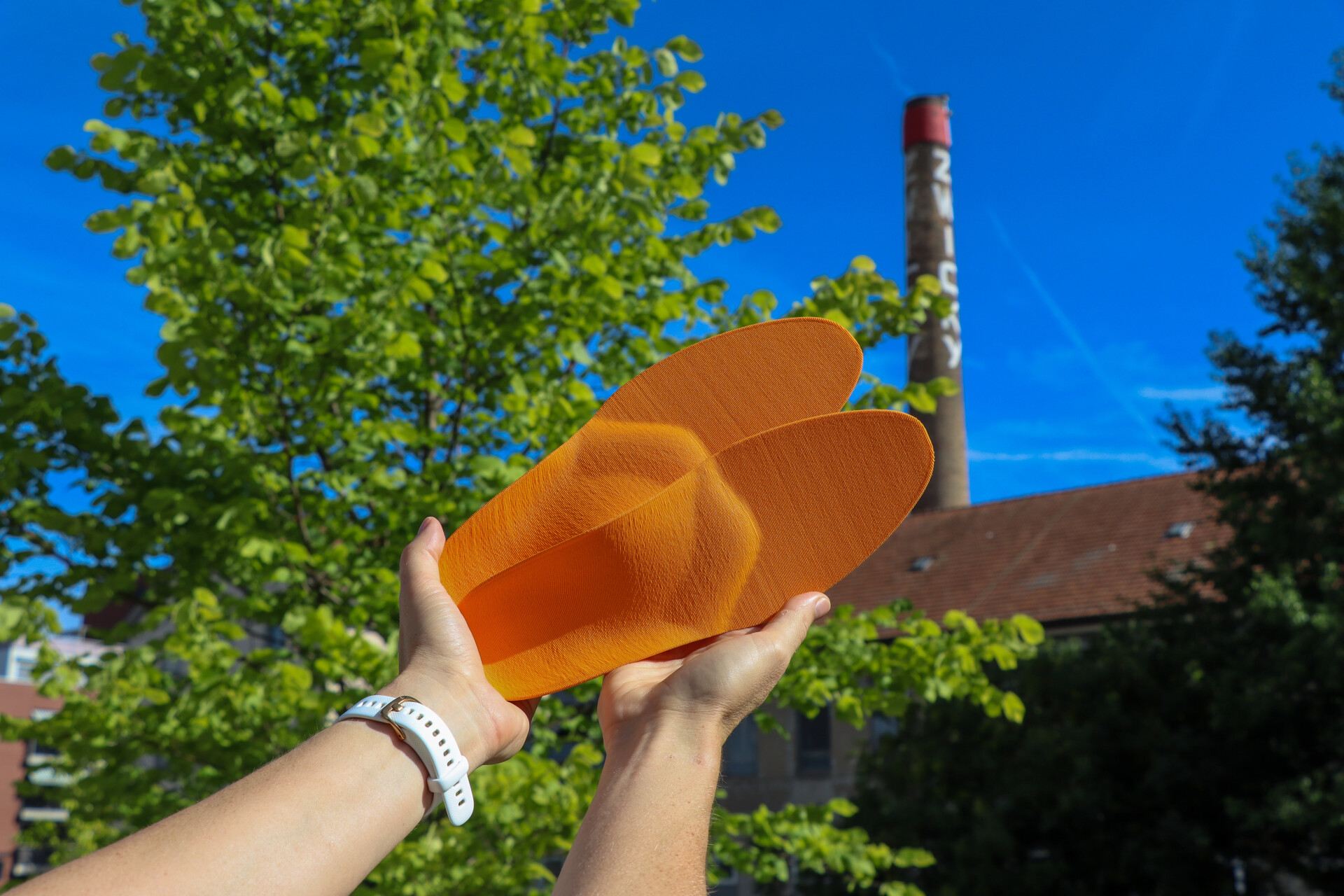 H&auml;nde halten ein Paar orange Support-Schuheinlagen, personalisiert nach 2D-Druckmessung, von Laufgsund vor dem markanten Zwicky-Schornstein in D&uuml;bendorf.