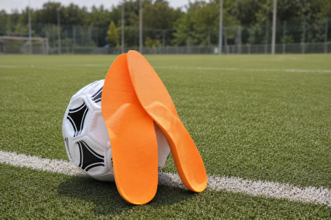 Zwei orange Support-Einlagen von Laufgsund lehnen an einem Fussball auf dem Rasenplatz &ndash; pr&auml;ventive Unterst&uuml;tzung und maximaler Komfort f&uuml;r gesunde F&uuml;sse beim Sport.