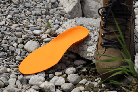 Eine orange Support-Einlage von Laufgsund liegt auf Kieselsteinen neben einem braunen Wanderschuh &ndash; f&uuml;r optimale Fussstabilit&auml;t und Halt beim Wandern im Gel&auml;nde.