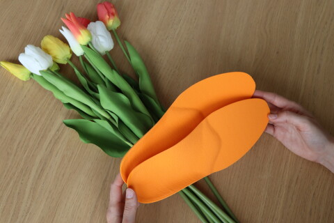 H&auml;nde halten zwei personalisierte, ergonomische orange Support-Einlagen von Laufgsund, platziert vor einem bunten Fr&uuml;hlingsstrauss aus Tulpen auf einem Holztisch.